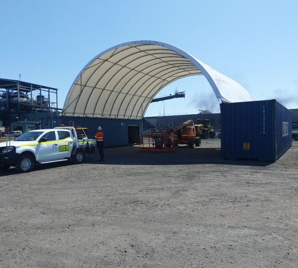 Shipping container canopy shelter spanning the gap between two blue shipping containers on an industrial site — covered equipment bay with drive-through access